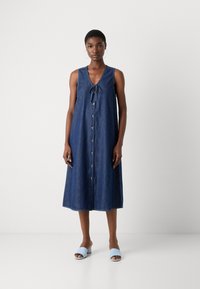 Mørkeblå denim ermeløs kjole med v-hals, knyting og knappestolpe foran. Brukt med lyseblå åpen tå mules.