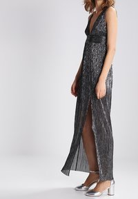 Metallic black long dress with a deep V-neck, textured vertical stripes, thigh-high slit, and fitted waist, paired with silver block-heel shoes.