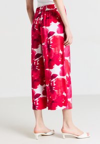 Une personne portant des pantalons à larges jambes cropped à motifs floraux rouges et blancs et des sandales à talons blancs, se tenant sur un sol clair avec un fond blanc.