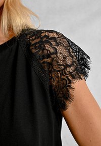 Top nero con maniche corte, rifinito con pizzo nero e bordi frangiati. Tessuto morbido con una vestibilità comoda.