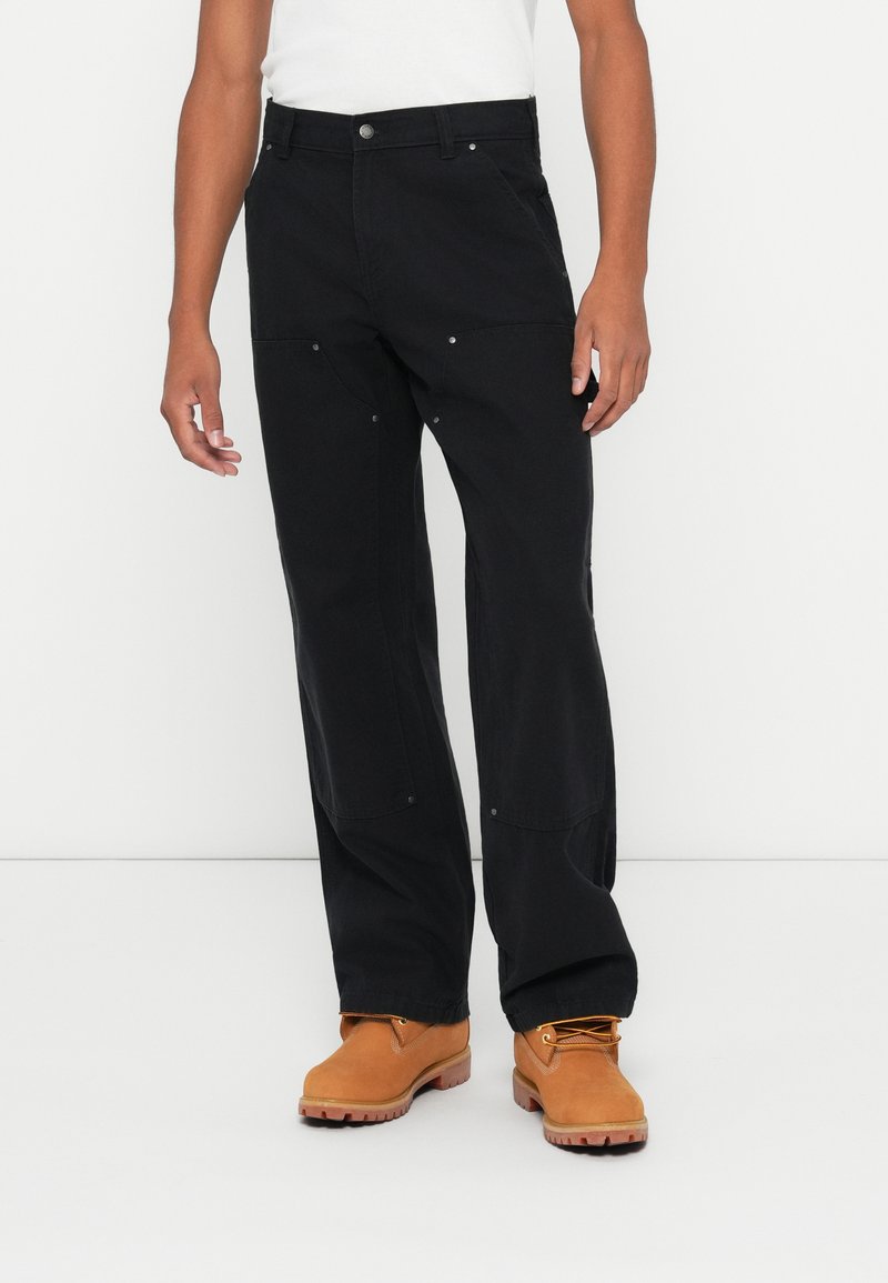Pantalons noirs à coupe ample avec poches avant et accents en métal argenté. Fabriqués à partir d'un tissu durable, associés à des bottes de travail couleur beige.