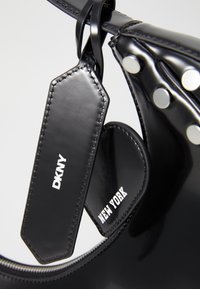 Sac en cuir verni noir orné de clous ronds argentés, avec deux étiquettes rectangulaires affichant "DKNY" et "NEW YORK" en texte blanc.