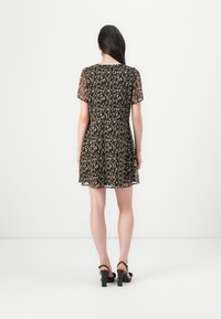 Femme aux longs cheveux foncés portant une robe à manches courtes noire et beige à motifs et des sandales noires à talons hauts, debout de dos.