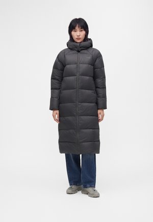 Kvinde iført en lang sort polstret vinterjakke med høj krave, blå jeans og grå sneakers, stående mod en enkel lys baggrund.