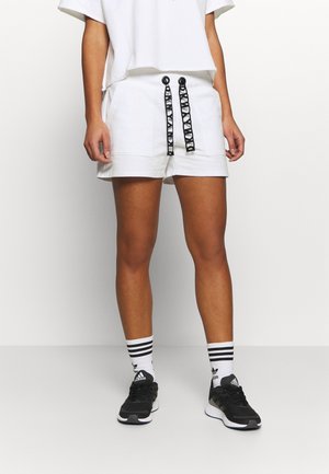 Osoba ubrana w białe szorty z czarnymi sznurkami w wzory, czarne trampki i białe skarpetki z czarnymi paskami, stojąca na tle jednolitej ściany.