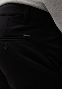 Pantalon noir sur mesure avec une poche arrière boutonnée et une petite étiquette de marque en métal près de la poche, porté avec un haut gris texturé.