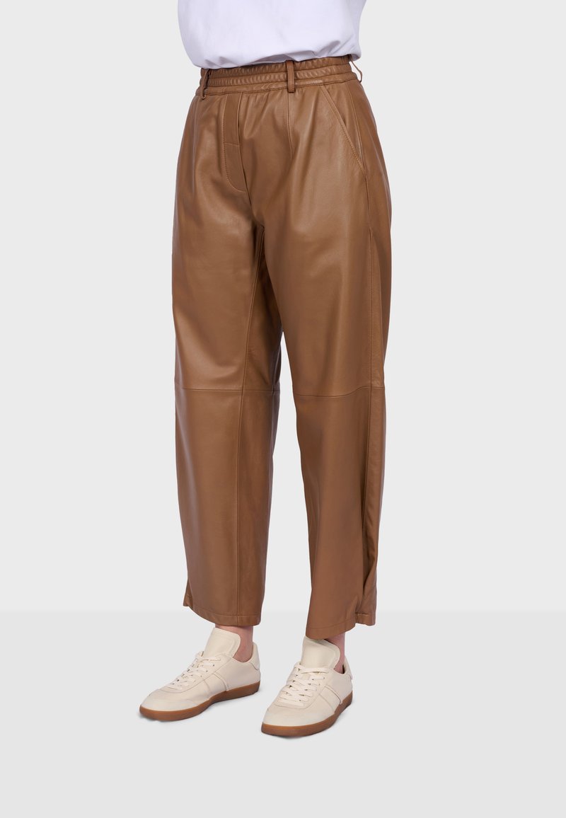 Braune High-Waist-Hose aus Kunstleder mit geraden Beinen, kombiniert mit weißen Turnschuhen, dargestellt an einer stehenden Person vor einem schlichten Hintergrund.