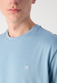 Mėlyna medvilninė marškinėliai su minkšta tekstūra ir apykakle, turi mažą baltą siuvinėtą "cK" logotipą kairiajame krūtinės plote.