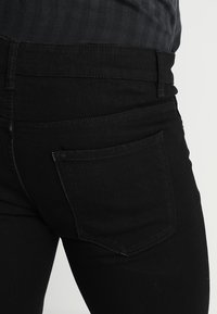 Primer plano de una persona que lleva vaqueros de mezclilla negros con un bolsillo trasero, combinados con una camiseta gris oscuro metida por dentro.