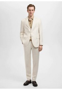 Costum de culoare deschisă, fabricat dintr-un material texturat, având o croială slim fit cu rever în v. Acesta este asortat cu o cămașă bej și mocasini închiși la culoare.
