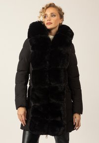 Frau in einem schwarzen gefütterten Mantel mit großen schwarzen Kunstfell-Elementen auf der Vorderseite, die vor einem hellen Hintergrund steht.