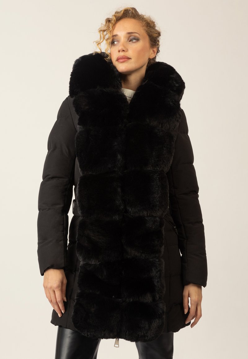 Frau in einem schwarzen gefütterten Mantel mit großen schwarzen Kunstfell-Elementen auf der Vorderseite, die vor einem hellen Hintergrund steht.