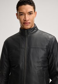 Schwarze Lederjacke mit hohem Kragen, Frontreißverschluss und Seitentaschen. Hat eine glatte Textur und Nahtdetails über der Brust.