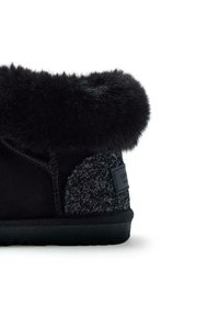 Bottine noire avec un revers en fourrure peluche et un patch de talon pailleté texturé, semelle plate et étiquette de marque à l'arrière.