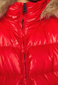 Doudoune rouge brillante avec fermeture éclair noire et bordure en fausse fourrure marron sur le col, montrée en gros plan sur la partie supérieure de la poitrine et le cou.