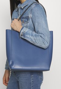 Niebieska syntetyczna torba typu tote o wyraźnym kształcie, gładkiej matowej fakturze i krótkim czarnym uchwycie, trzymana na jasnym tle.