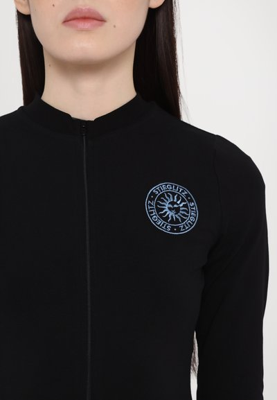 Chaqueta negra con cremallera y cuello alto, con un logo circular azul bordado que presenta un diseño de sol en el lado izquierdo del pecho.