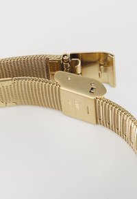 Bracelet de montre en maille en acier inoxydable doré avec boucle de fermeture à ouverture sur fond blanc.