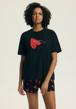 Kobieta w czarnej, oversize'owej koszulce z czerwonym sercem i napisem, w połączeniu z czarnymi szortami w czerwony wzór serc.