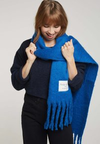 Écharpe tricotée bleue en matériau doux, avec des bords frangés et une étiquette cousue. Le design est large et s'enroule confortablement autour du cou.