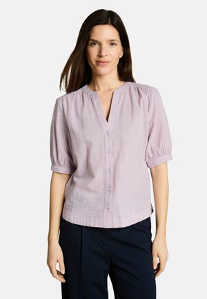 Kobieta ubrana w jasnofioletową, krótką koszulę na guziki z lekko bufkami na rękawach oraz czarne spodnie, stojąca na białym tle.