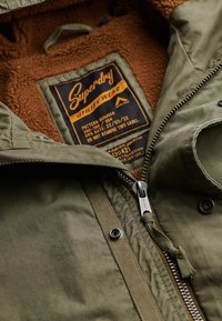 Parka de utilidad verde con un forro de felpa marrón, cierre frontal y etiqueta de marca. Presenta un tejido duradero y elementos ajustables.