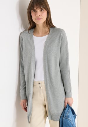 Donna che indossa un cardigan grigio chiaro aperto sul davanti, camicia bianca, pantaloni beige, con in mano una giacca di jeans blu piegata contro una parete bianca.