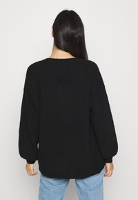 Kvinna med långt mörkt hår iförd en löst sittande svart stickad tröja och ljusblå jeans, vänd bort från kameran mot en enkel vit bakgrund.