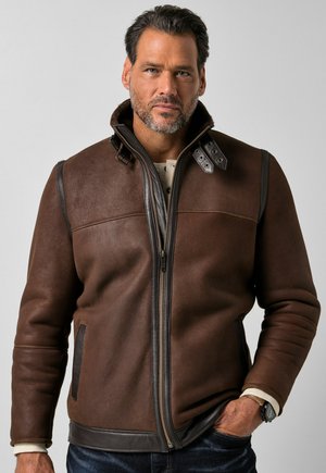 Homme aux cheveux courts et foncés et à la barbe, portant une veste en cuir marron zippée avec un col haut, debout avec une main dans la poche devant un fond uni.
