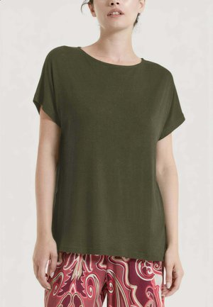 Donna che indossa una maglietta verde oliva a maniche corte abbinata a pantaloni rossi con motivo paisley, in piedi su uno sfondo chiaro.