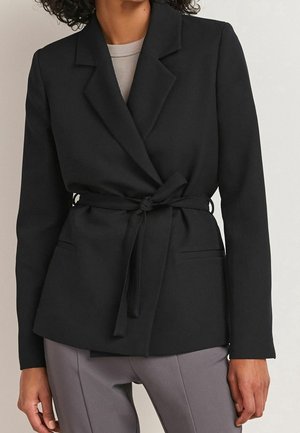 Femme portant un blazer noir ceinturé sur un haut beige et un pantalon gris, debout devant un fond uni de couleur claire.