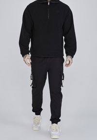 Czarna bluza polarowa na zamek z wysokim kołnierzem, zestawiona z czarnymi spodniami cargo z bocznymi kieszeniami i sznureczkowymi detalami oraz białymi trampkami.