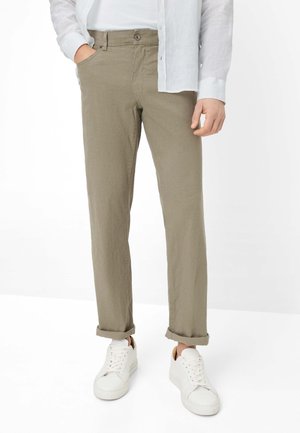 Uomo con pantaloni beige risvoltati, sneaker bianche e camicia bianca a maniche lunghe, con una mano in tasca.