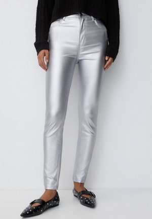 Bas du corps d'une personne portant un pantalon slim métallique argenté, un pull noir et des ballerines noires ornées de clous argentés, sur fond blanc.