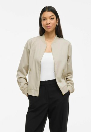 Jeune femme aux cheveux longs portant une veste bomber beige, un haut blanc et un pantalon noir, se tenant les mains dans les poches devant un fond blanc.