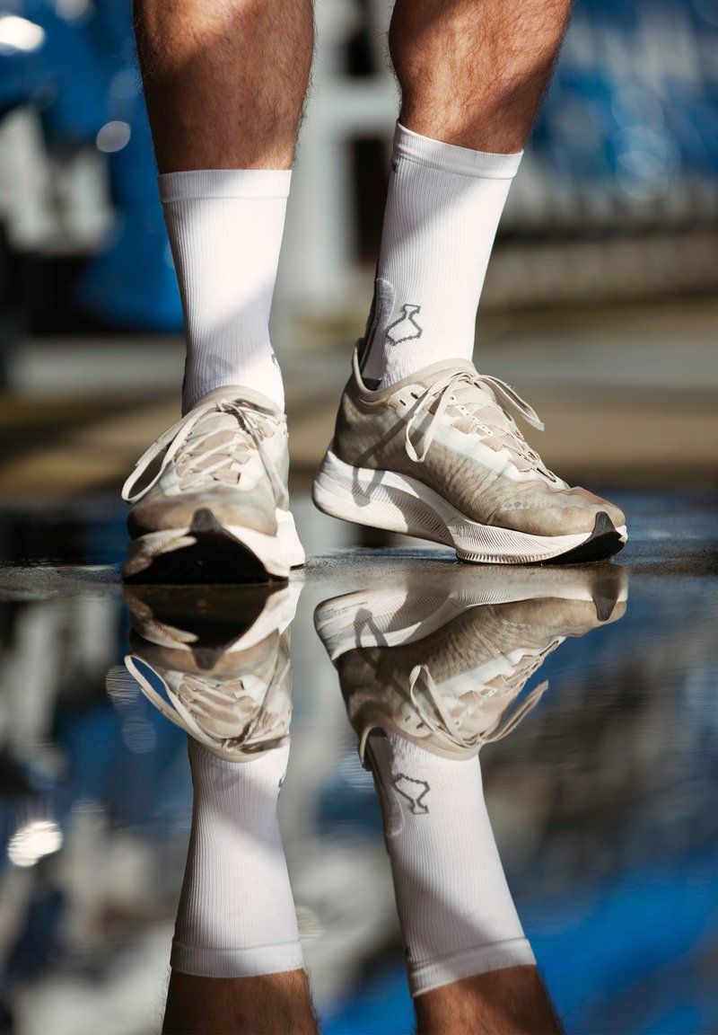 Jasno beżowe buty sportowe z teksturowaną cholewką i białymi akcentami, w połączeniu z białymi skarpetkami za kostkę. Odbicie widoczne na mokrej powierzchni.