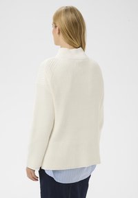 Femme aux cheveux blonds portant un pull en maille blanche sur une chemise rayée bleue et un pantalon sombre, de dos contre un fond uni.
