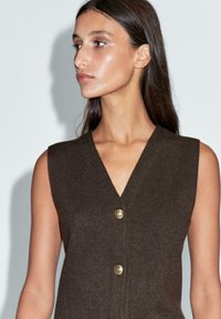 Robe marron sans manches avec un décolleté en V et deux boutons dorés, réalisée en tissu texturé avec une silhouette ajustée.