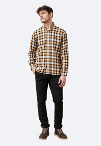 Camisa de cuadros en negro, amarillo y blanco, con cuello abotonado y bolsillo en el pecho; combinada con pantalones ajustados negros y botas marrones.