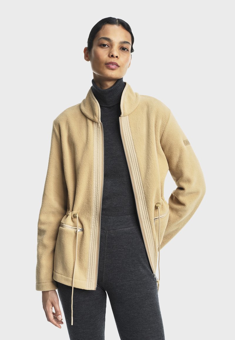 Veste en polaire beige avec un col haut, fermeture éclair à l'avant et taille ajustable par cordon. Inclut deux poches latérales et un tissu texturé.