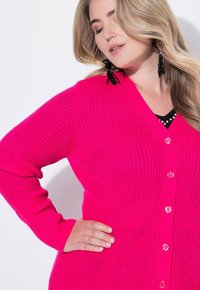 Kobieta z długimi blond włosami, nosząca czarne koralikowe kolczyki i jasnoróżowy sweter zapinany na guziki, z ręką opartą na biodrze.