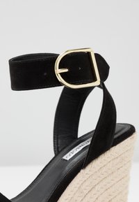 Sandale en daim noir avec une boucle dorée et une semelle compensée en jute tissé, dotée d'une semelle intérieure en cuir lisse et d'une bride à la cheville.