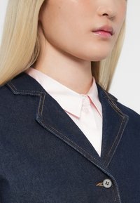 Traper jakna s izrezanim ovratnikom i vidljivim narančastim šavovima, složena preko svijetloružaste košulje na dugmad, koja prikazuje elegantan dizajn.