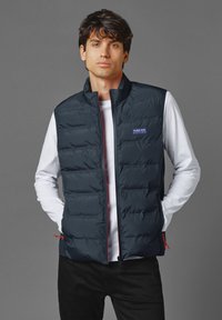 Marinblå pufferväst med hög krage, lättviktig, kviltad design, med röda dragkedjor och en Red Bull Racing-logotyp på bröstet.