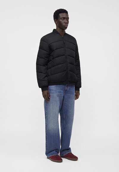 Homme debout portant une veste matelassée noire, un jean bleu ample et des baskets rouges sur un fond clair uni.