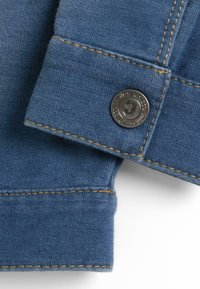 Tissu en denim bleu avec une couture visible et des points de surfilage jaunes contrastants ; présente un bouton en argent portant le nom de la marque gravé dessus.