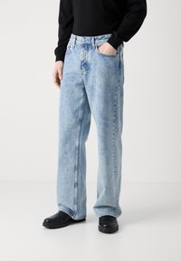 Light blue, wide-leg denim jeans with a faded finish, five pockets, and a button closure. Worn with black clogs, textured surface visible.