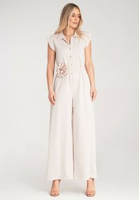 Jasno beżowy kombinezon z krótkimi rękawami, z guzikami z przodu oraz kwiatowym akcentem w talii. Wykonany z miękkiego, gładkiego materiału z szerokimi nogawkami.