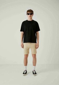 Black cotton T-shirt and beige shorts, paired with black sneakers, white socks, and sunglasses, standing against a neutral background.