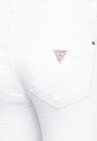 Jeans de mezclilla blanca con un parche en la parte trasera del bolsillo que presenta un logo triangular de "Guess" en rojo y blanco. Textura suave con costuras visibles.